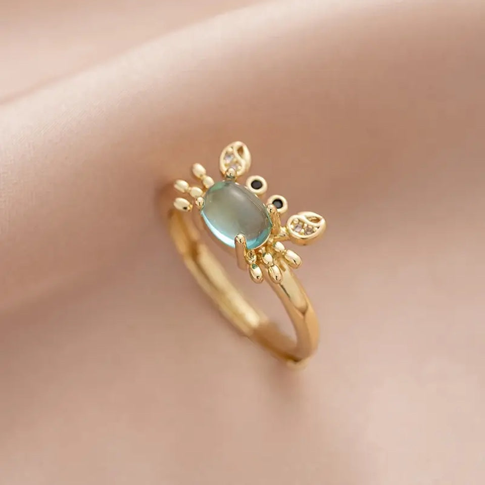 une superbe bague dorée réglable en forme de crabe qui lève les pinces. Son corps est un joli strass bleu transparent qui rappelle la mer. Ses pinces ont des strass.