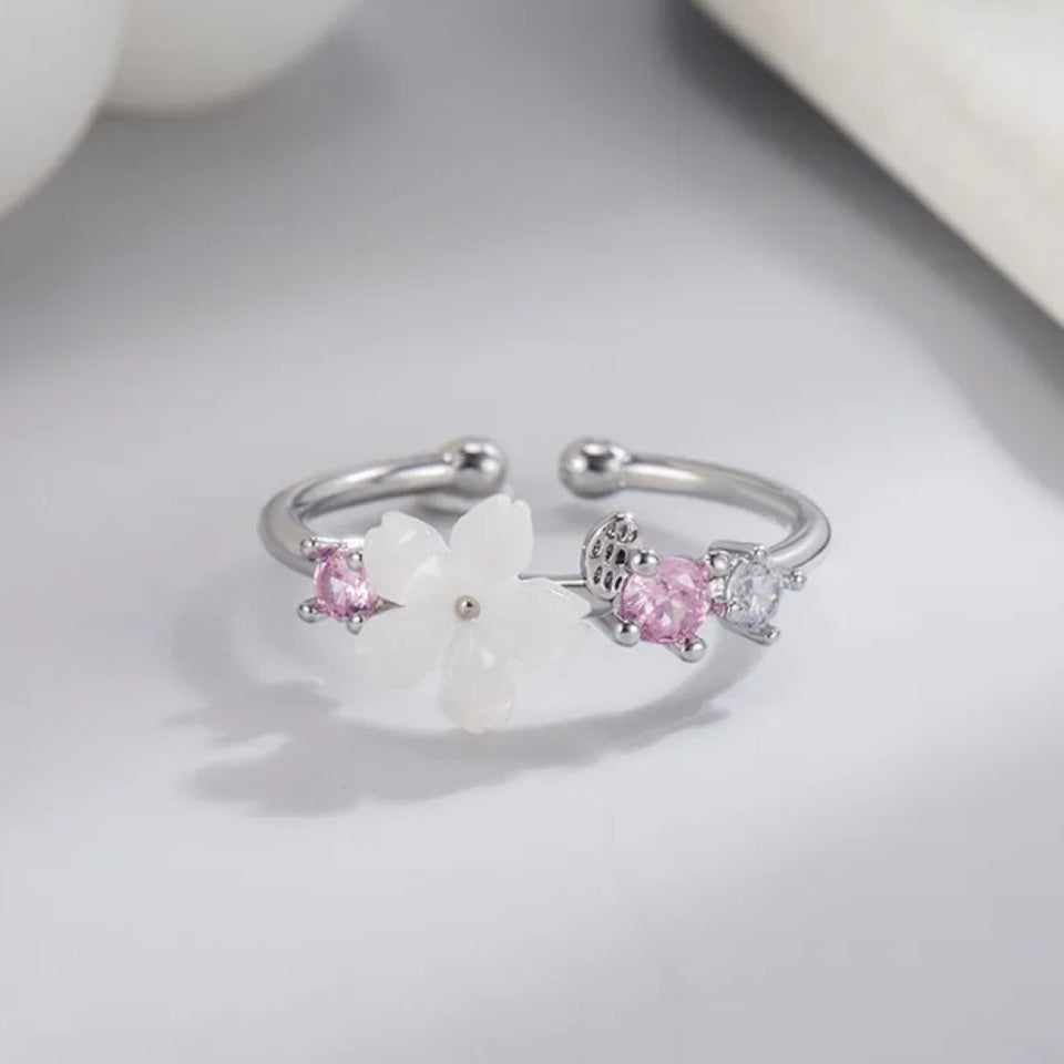 une superbe bague argentée et réglable d'une fleur de sakura blanche accompagnée de petits strass roses et blancs.