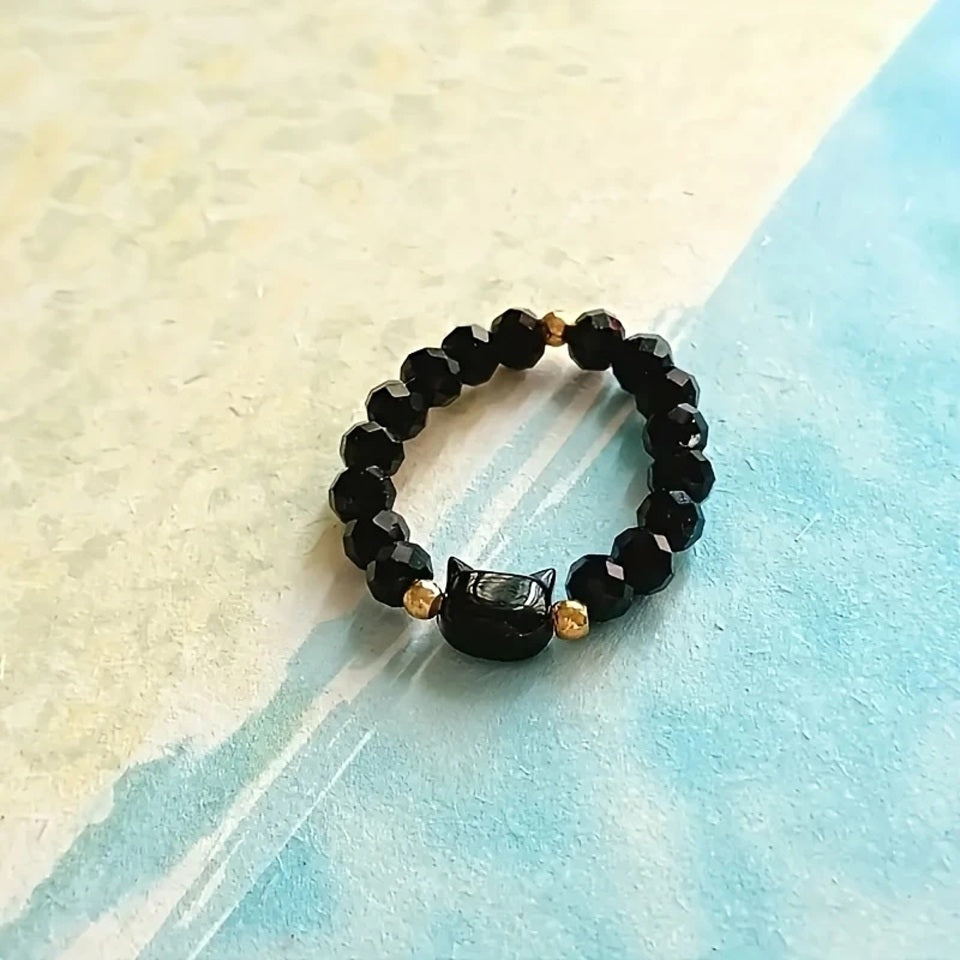 une bague taille élastique toute en parles et une breloque perles en forme de tête de chat. Disponible en coloris noir ou blanc.