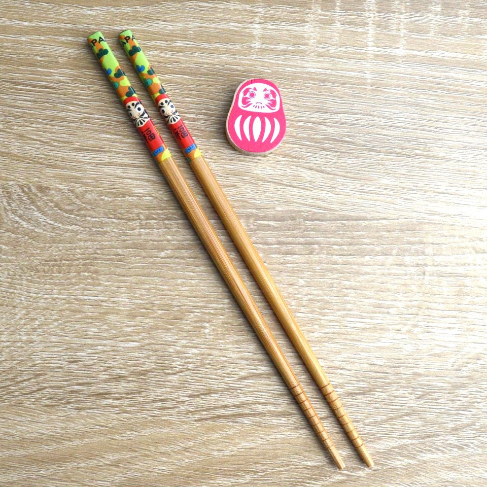 une paire de baguettes japonaises décorées d'un daruma rouge avec son repose baguette en forme de daruma. Folklore et style se combinent pour une jolie table inspiré du Japon.