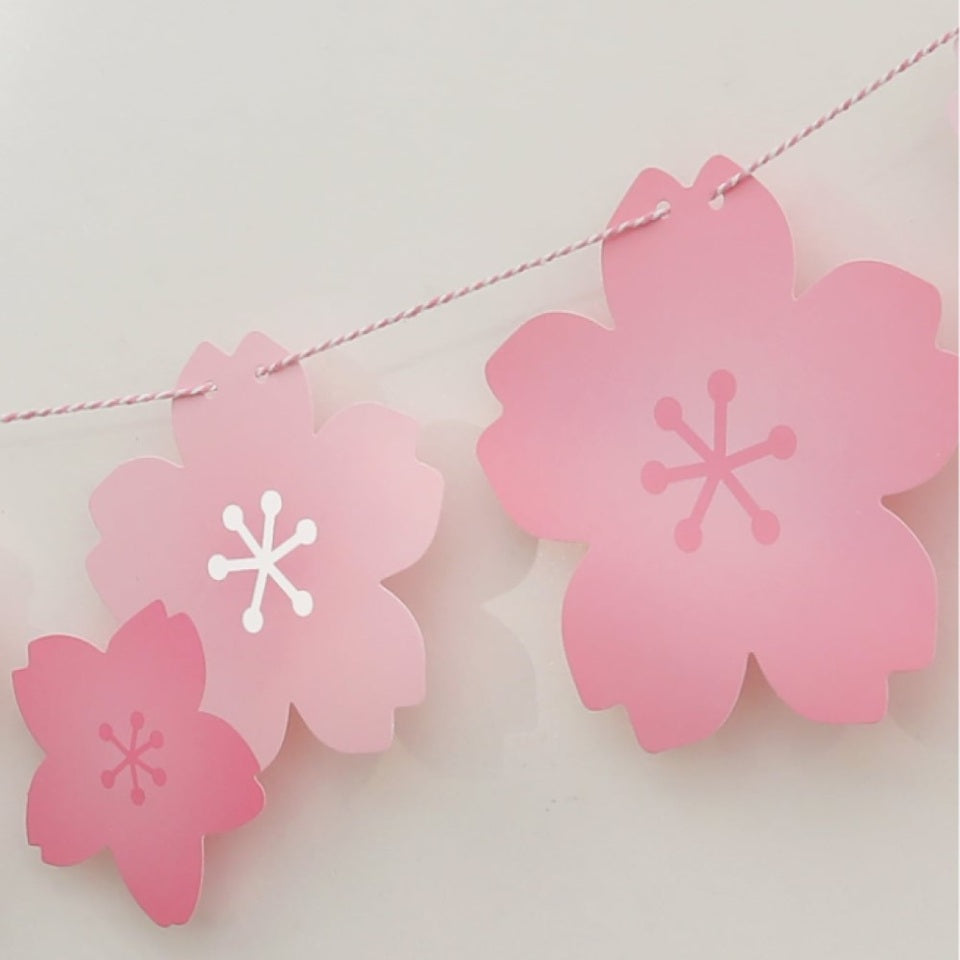 une banderole en papier formée de fleurs de sakura rose. A suspendre avec sa cordelette fournie. Elle ajoute une touche florale et mignonne à une pièce ou un extérieur.