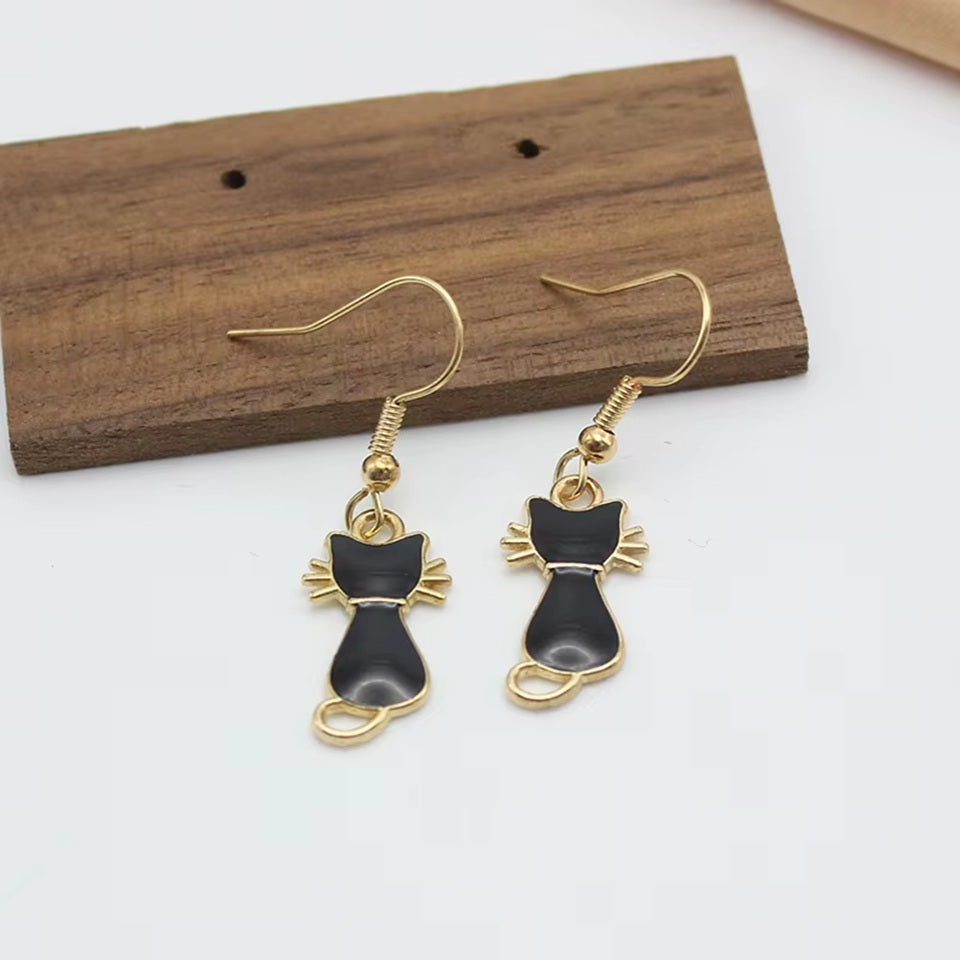 une paire de boucles d'oreilles dorées en forme de chats noirs, on voit leurs petites moustaches qui dépassent adorablement. Look mignon et tendre !