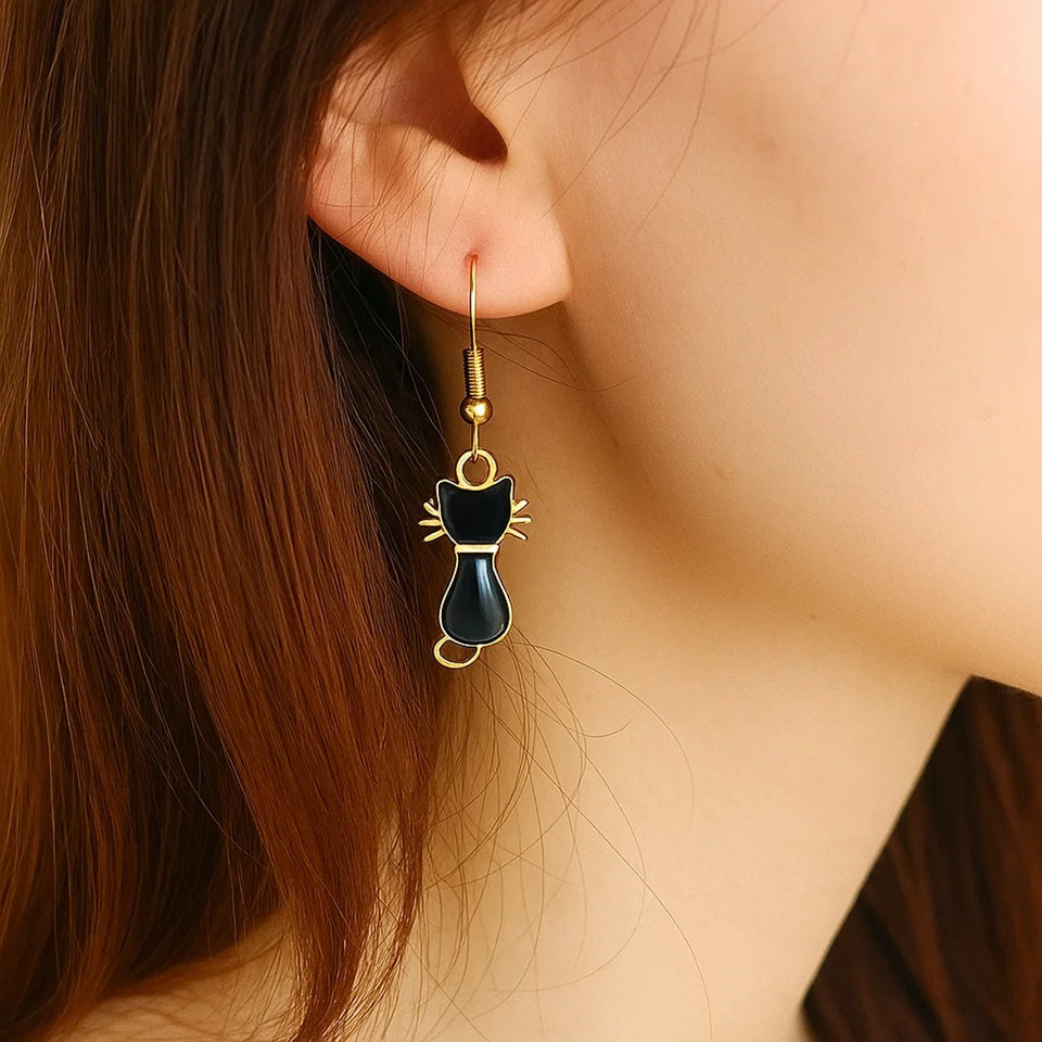 une paire de boucles d'oreilles dorées en forme de chats noirs, on voit leurs petites moustaches qui dépassent adorablement. Look mignon et tendre !