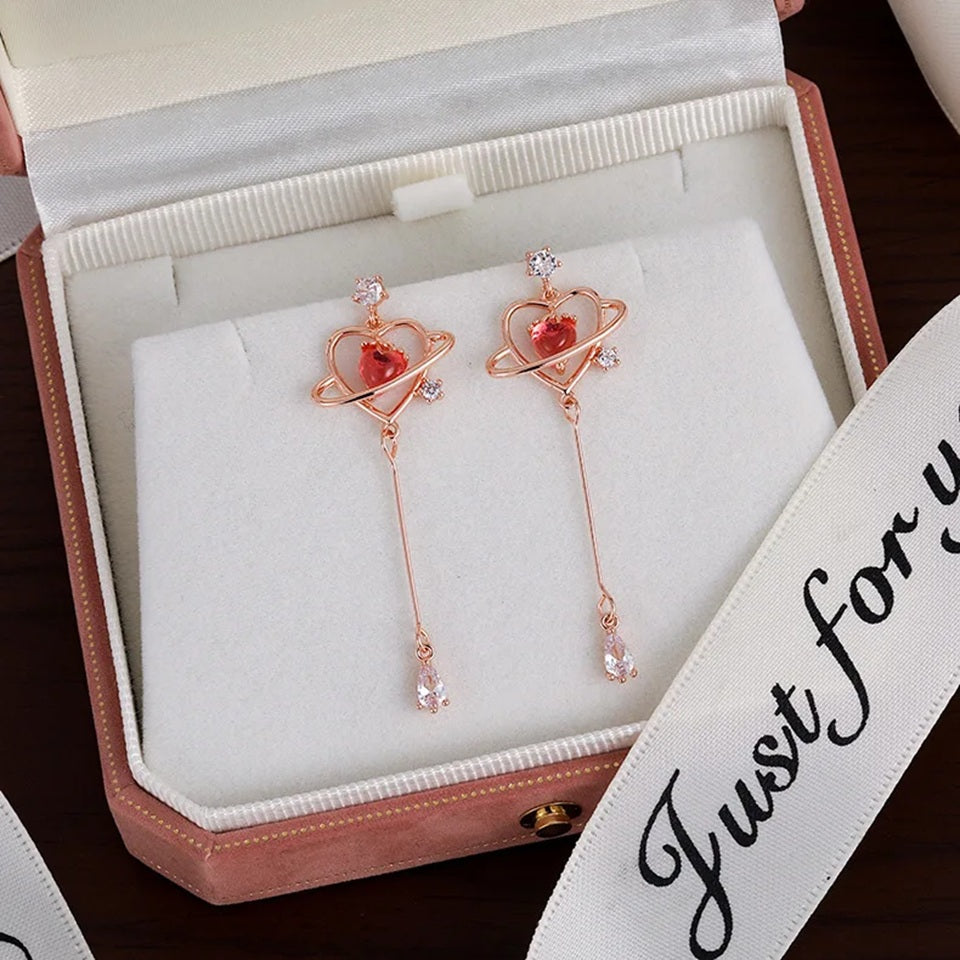 des boucles d'oreilles au design romantique et féminin : une structure en forme de coeur doré abrite une strass rose en forme de coeur, tel le coeur d'une planète. Au bout, est suspendu un strass brillant en forme de goutte.