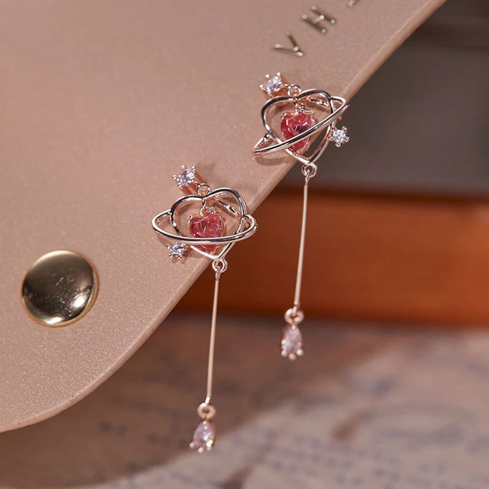 des boucles d'oreilles au design romantique et féminin : une structure en forme de coeur doré abrite une strass rose en forme de coeur, tel le coeur d'une planète. Au bout, est suspendu un strass brillant en forme de goutte.