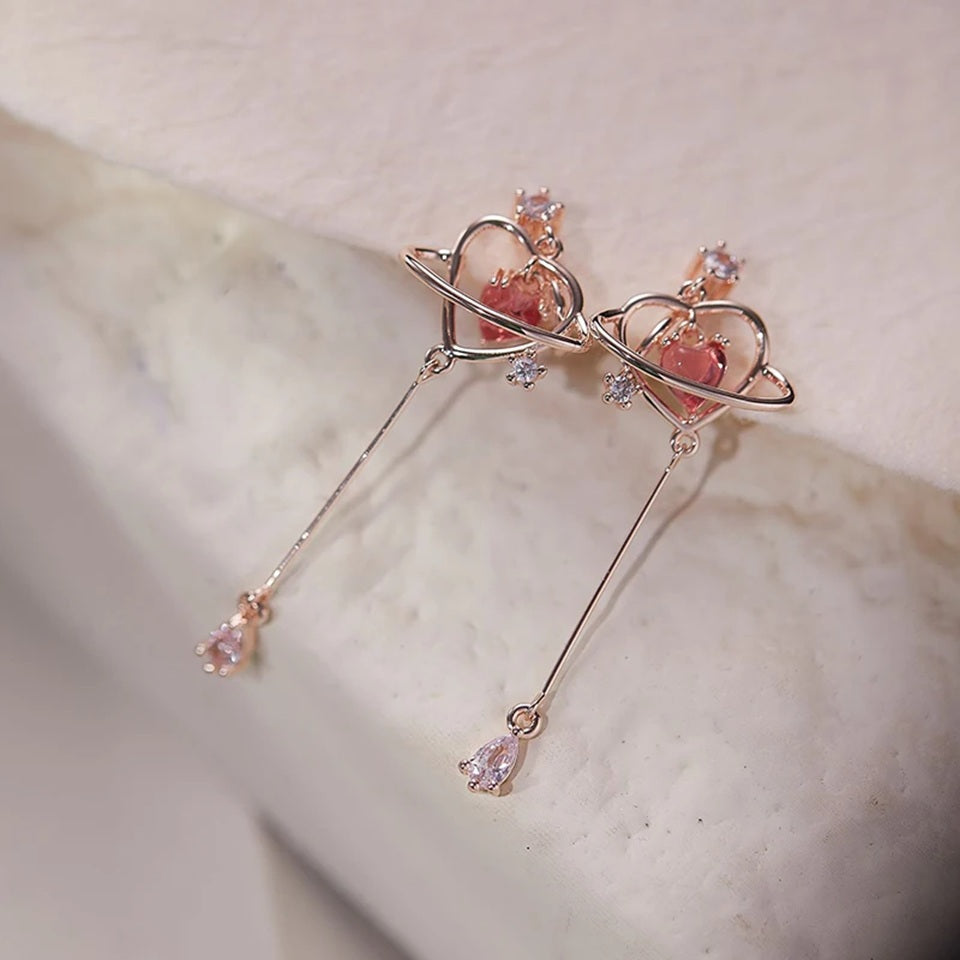 des boucles d'oreilles au design romantique et féminin : une structure en forme de coeur doré abrite une strass rose en forme de coeur, tel le coeur d'une planète. Au bout, est suspendu un strass brillant en forme de goutte.