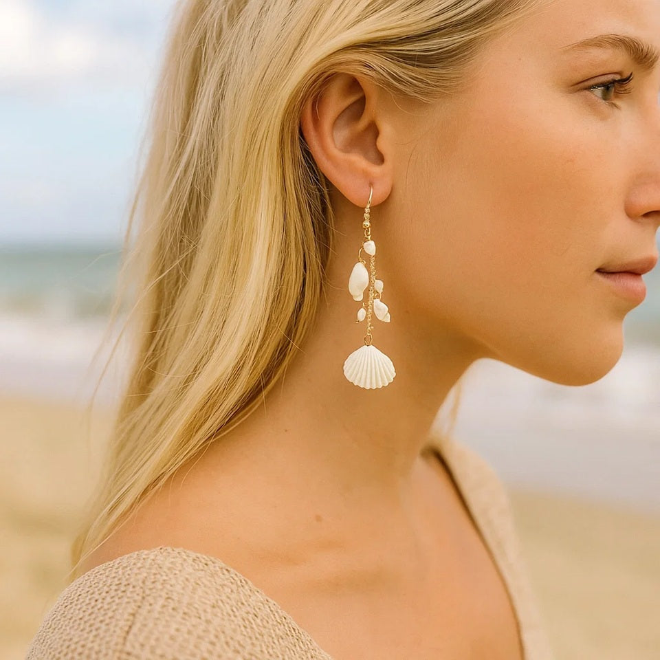 des boucles d'oreilles longues et dorées formées de divers coquillages blancs. L'ensemble évoque les vacances, la plage et l'été.