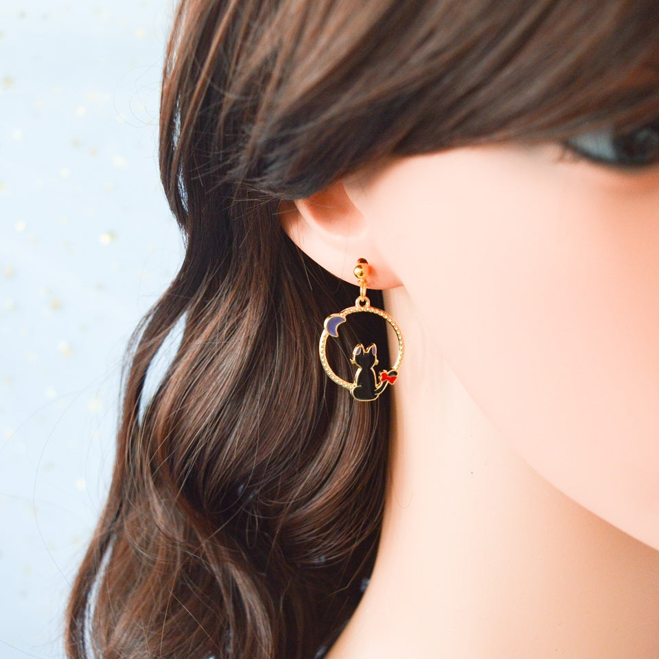une paire de boucles d'oreilles asymétriques inspirées de Kiki la petite sorcière. D'un côté il y a Jiji le chat noir et de l'autre, Kiki sur son balai.