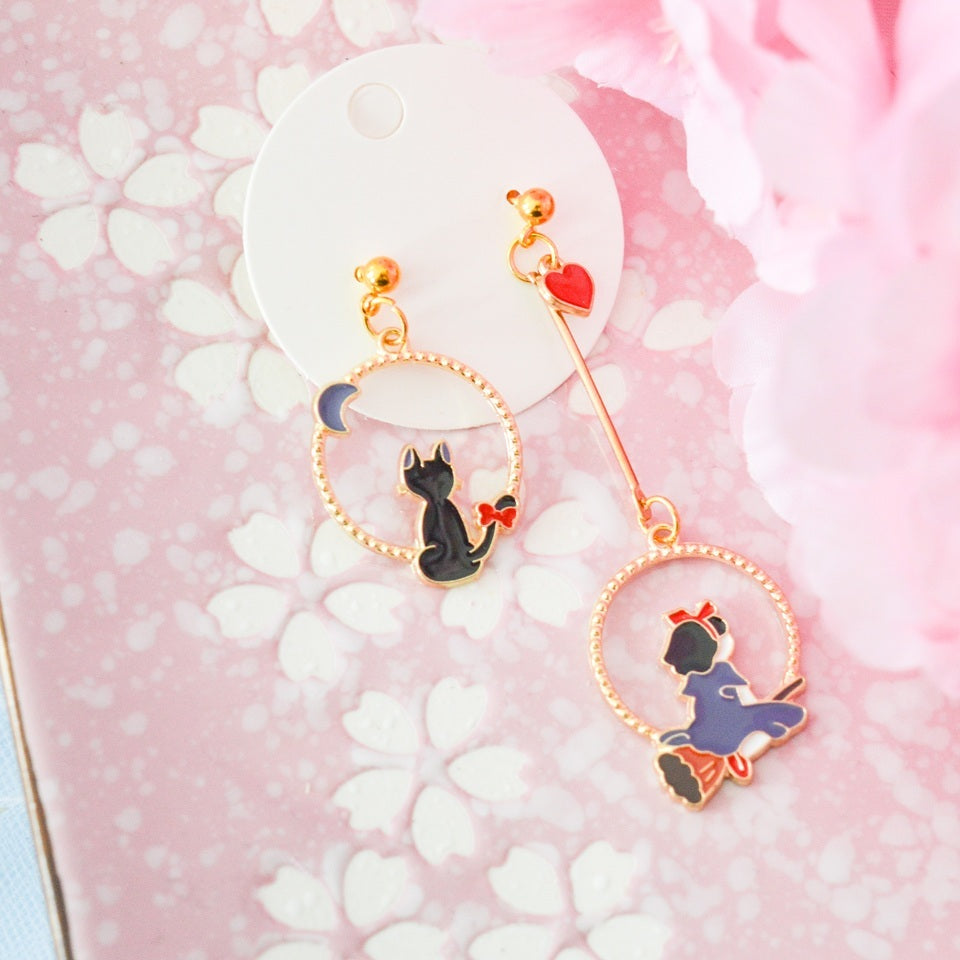une paire de boucles d'oreilles asymétriques inspirées de Kiki la petite sorcière. D'un côté il y a Jiji le chat noir et de l'autre, Kiki sur son balai.