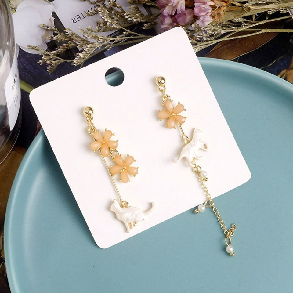 des boucles d'oreilles asymétriques et dorées. Un chat au ton nacré est posé tranquillement parmi des fleurs de sakura roses. L'ensemble est élégant et raffiné.