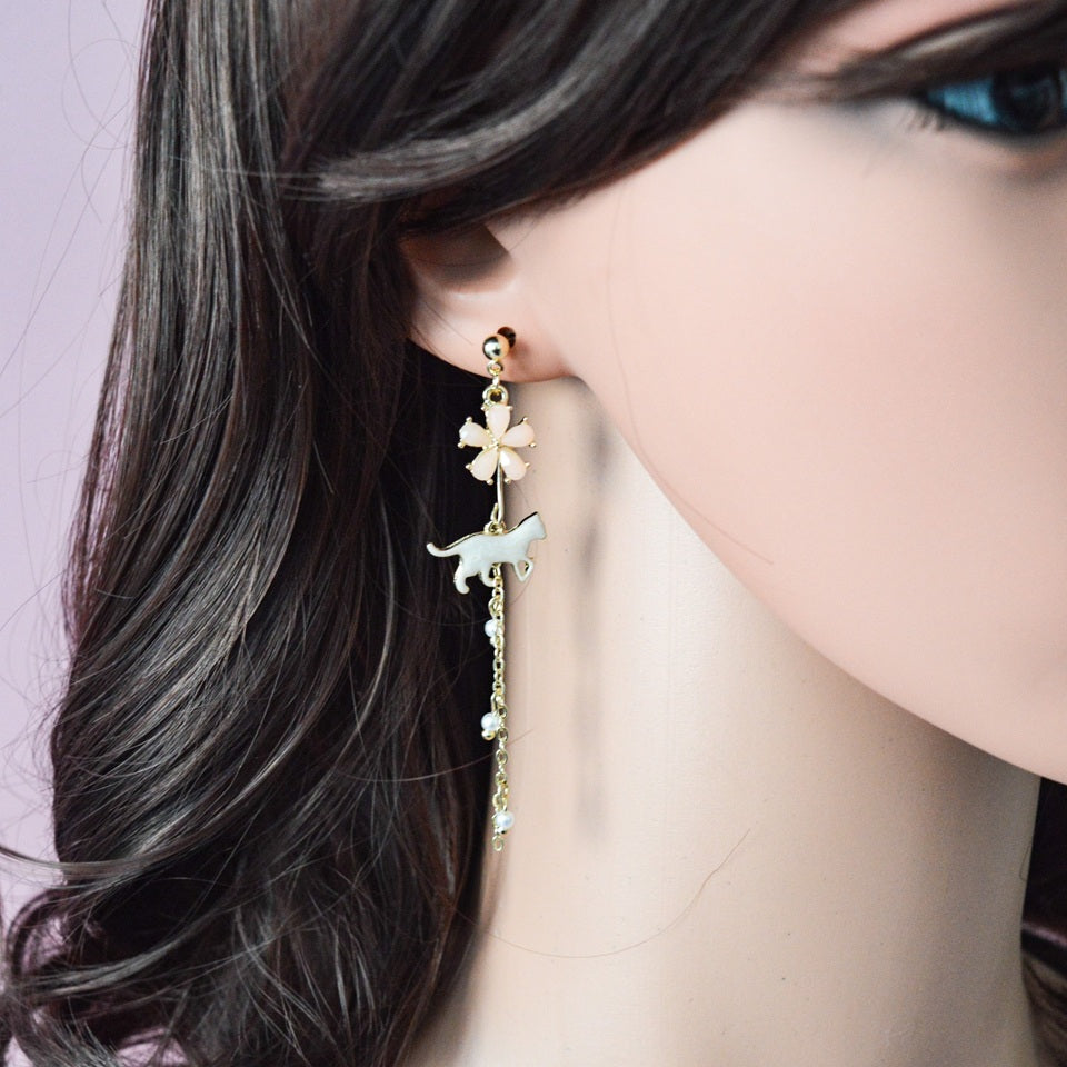 des boucles d'oreilles asymétriques et dorées. Un chat au ton nacré est posé tranquillement parmi des fleurs de sakura roses. L'ensemble est élégant et raffiné.