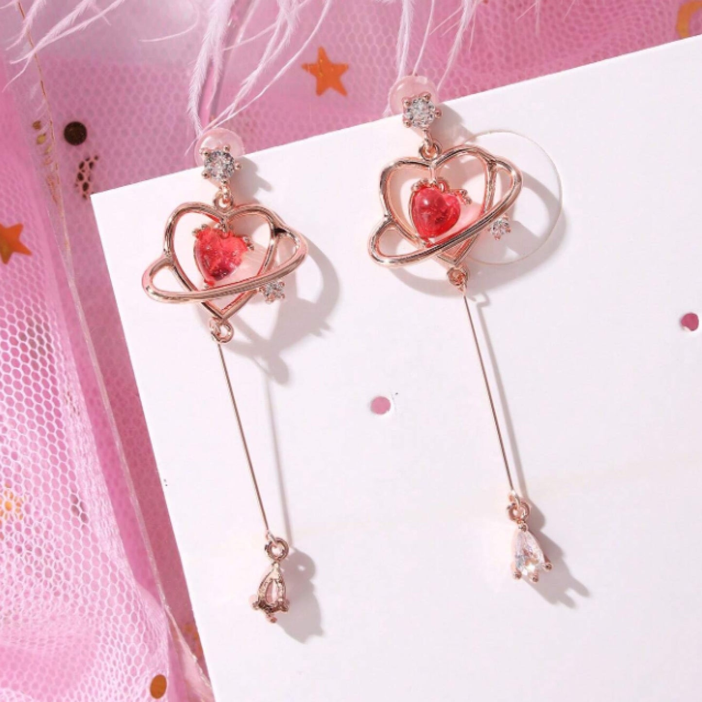 des boucles d'oreilles au design romantique et féminin : une structure en forme de coeur doré abrite une strass rose en forme de coeur, tel le coeur d'une planète. Au bout, est suspendu un strass brillant en forme de goutte.