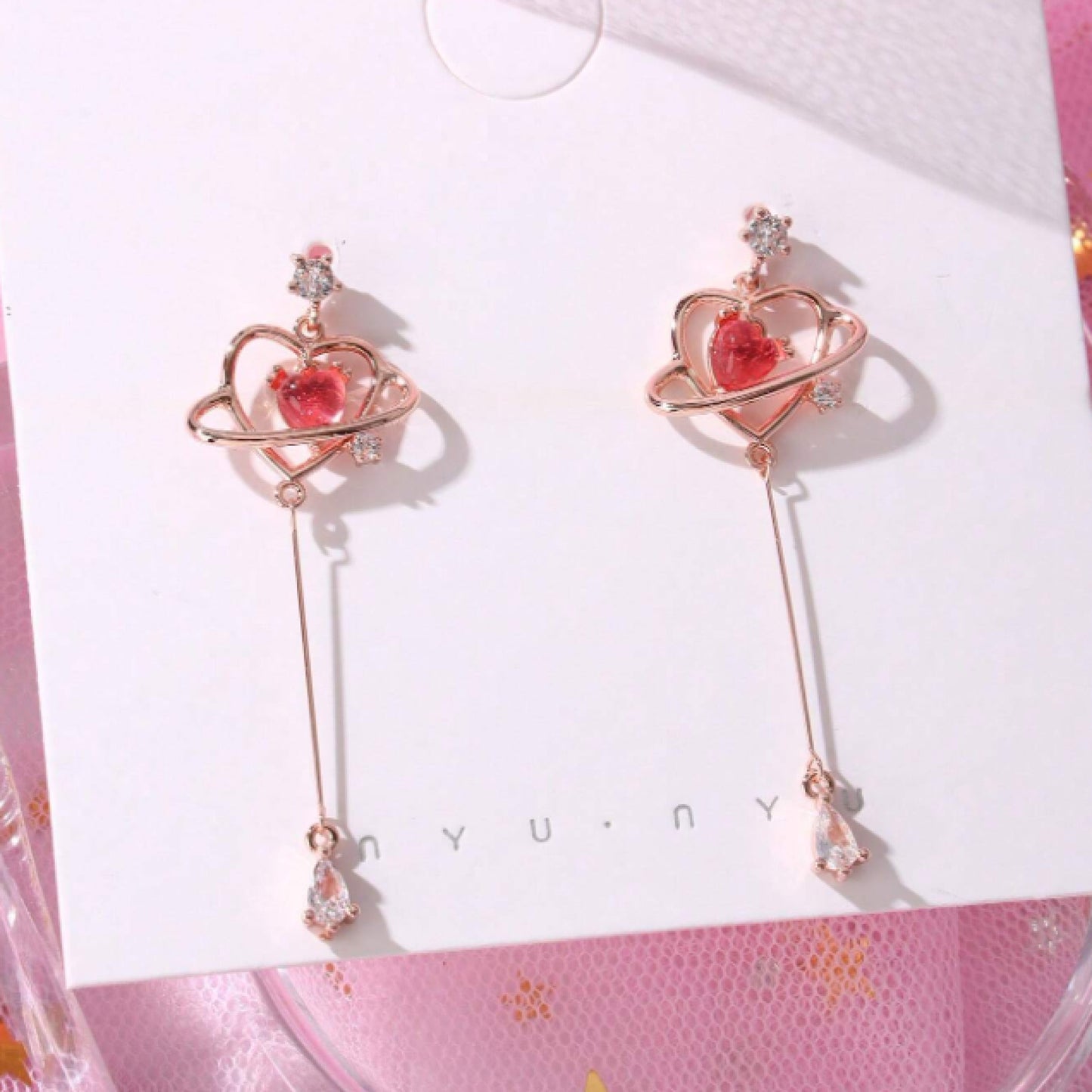 des boucles d'oreilles au design romantique et féminin : une structure en forme de coeur doré abrite une strass rose en forme de coeur, tel le coeur d'une planète. Au bout, est suspendu un strass brillant en forme de goutte.