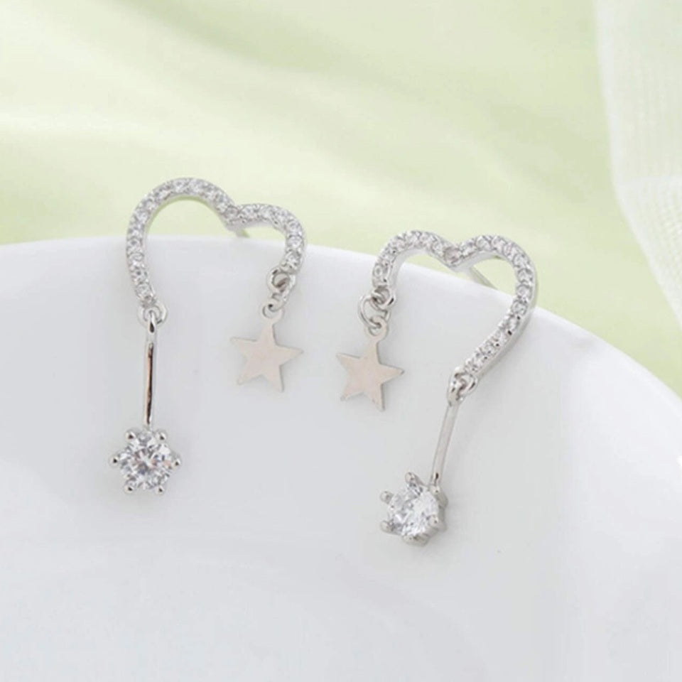 une paire de boucles d'oreilles en forme de coeur tout en strass. A chaque bout du coeur, est suspendu une étoile et un strass brillant.