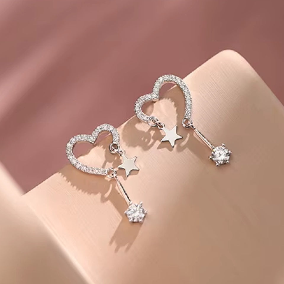 une paire de boucles d'oreilles en forme de coeur tout en strass. A chaque bout du coeur, est suspendu une étoile et un strass brillant.