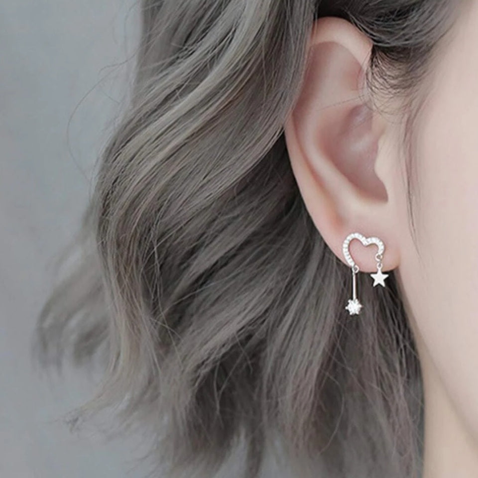 une paire de boucles d'oreilles en forme de coeur tout en strass. A chaque bout du coeur, est suspendu une étoile et un strass brillant.