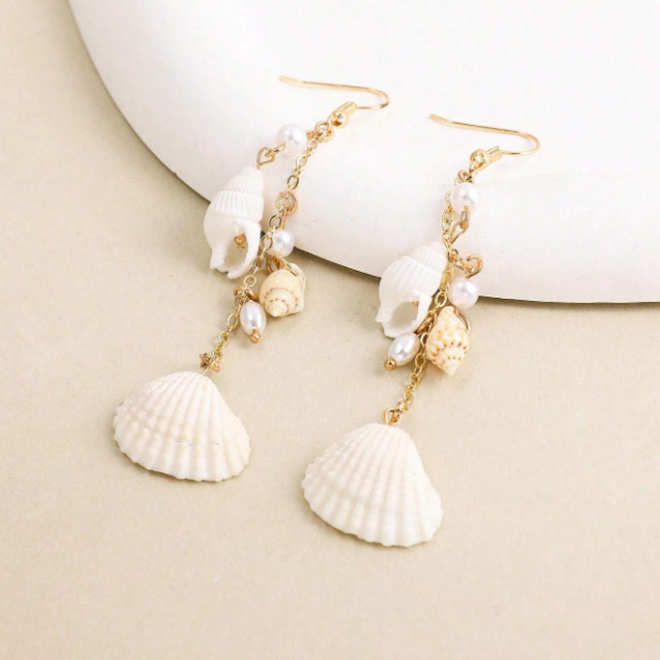 des boucles d'oreilles longues et dorées formées de divers coquillages blancs. L'ensemble évoque les vacances, la plage et l'été.