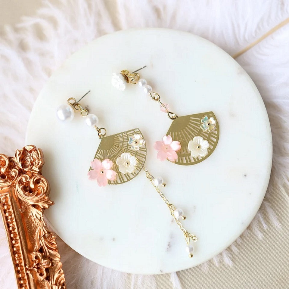 une paire de boucles d'oreilles asymétriques composées d'éventails dorés décorés avec de jolies fleurs de sakura, des perles blanches viennent apporter de l'élégance à l'ensemble.