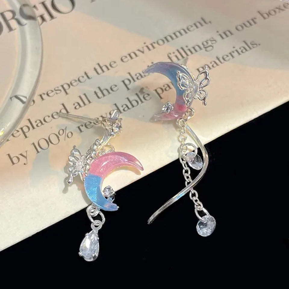 des boucles d'oreilles asymétriques en forme de lune bicolore bleue et rose sur laquelle est posé un papillon argenté. Une chainette y est suspendue au bout de laquelle pend un strass enforme de goutte.