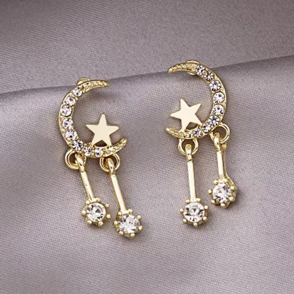 une paire de boucles d'oreilles en forme de lune toute en strass dans laquelle repose une étoile dorée. Deux strass sont suspendus à la lune et apportent encore plus de brillance.