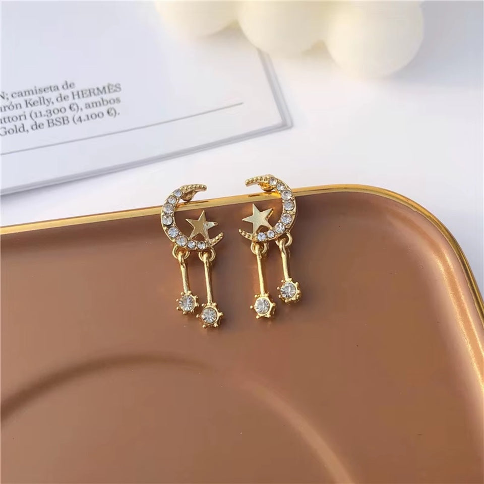 une paire de boucles d'oreilles en forme de lune toute en strass dans laquelle repose une étoile dorée. Deux strass sont suspendus à la lune et apportent encore plus de brillance.