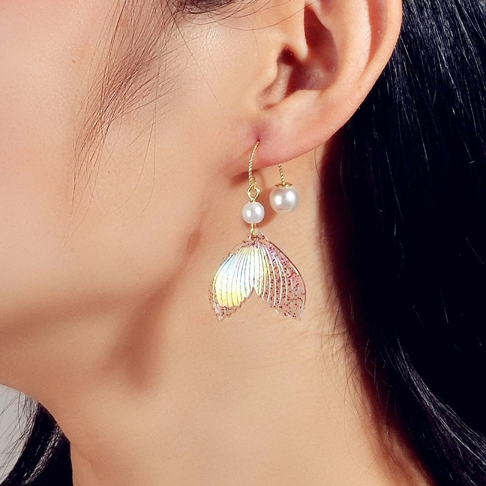 des boucles d'oreilles en forme de queues de sirènes pailletées et roses. De jolies perles blanches viennent ajouter de l'élégance à l'ensemble