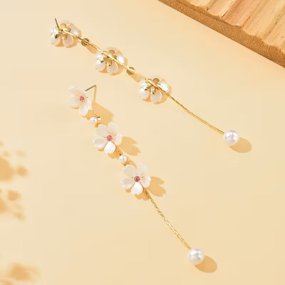 une superbe paire de boucles d'oreilles présentant trois fleurs de sakura blanches dont le coeur est un strass rose suspendues à une chaine dorée qui termine par une perle blanche. Le bijou est élégant, féminin et poétique.