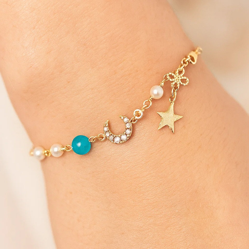 un bracelet doré élégant qui rappelle le mystère et la brillance de la galaxie. Il est composé d'un croissant de lune tout en strass, d'une étoile et de perles blanches et d'une bleue.