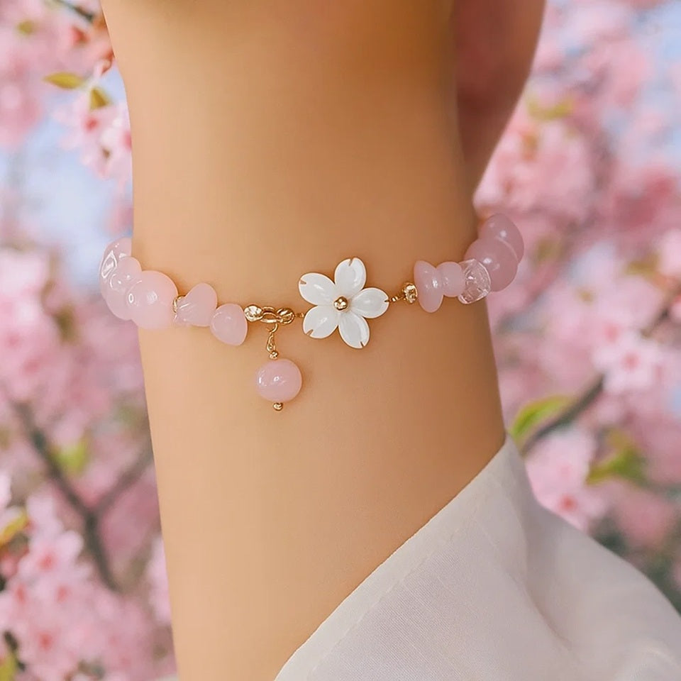 un bracelet doré composé de pierres et de perles roses pâles et d'une fleur de sakura. L'ensemble est raffiné et élégant.