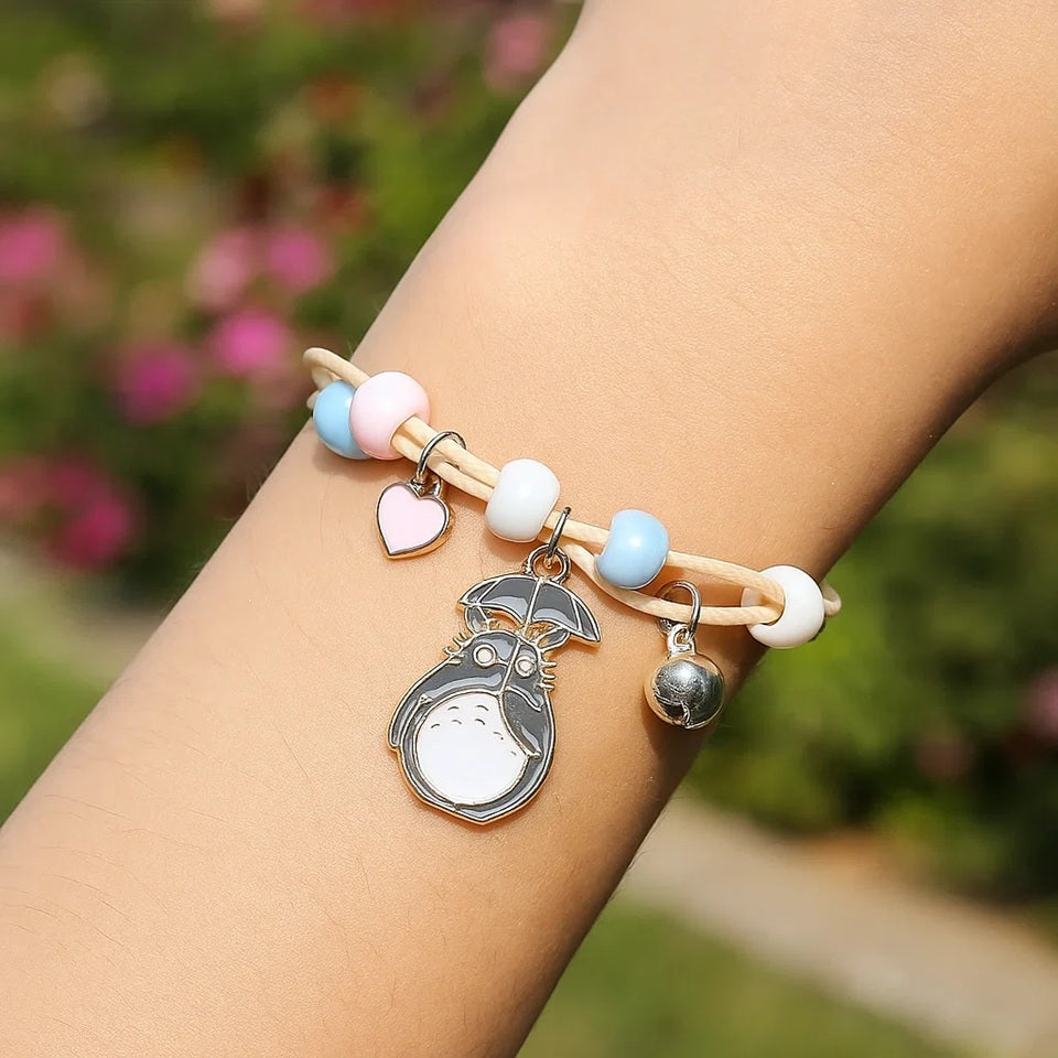 un bracelet en corde avec une breloque Totoro qui tient un parapluie. Sur le bracelet, il ya des perles bleu, rose et blanches avec un joli coeur rose et une clochette.