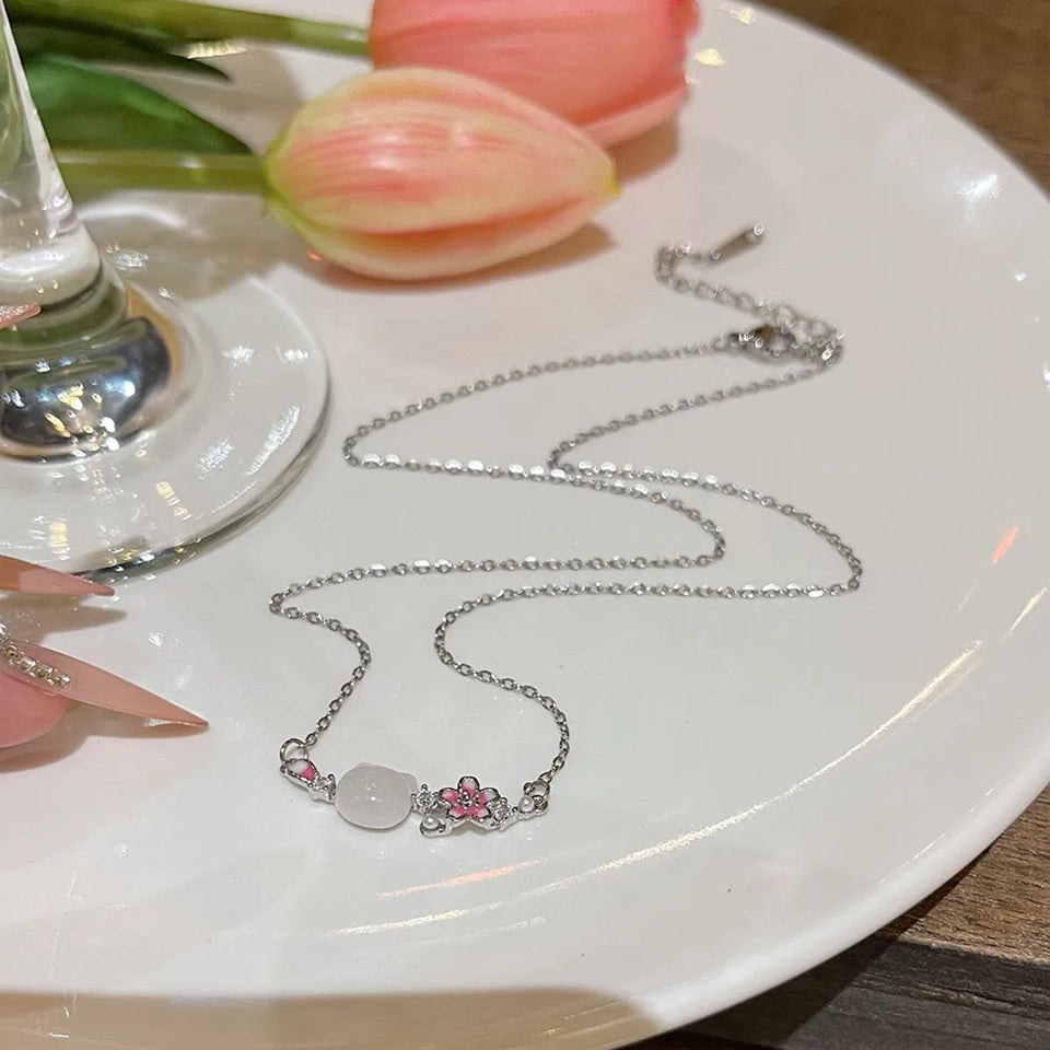 un superbe collier en acier inoxydable argenté composé d'une pierre blanche sculptée en forme de tête de chat, d'une fleur de sakura et d'un pétale rose et de petites perles blanches. L'ensemble est poétique, raffiné et plein de poésie.