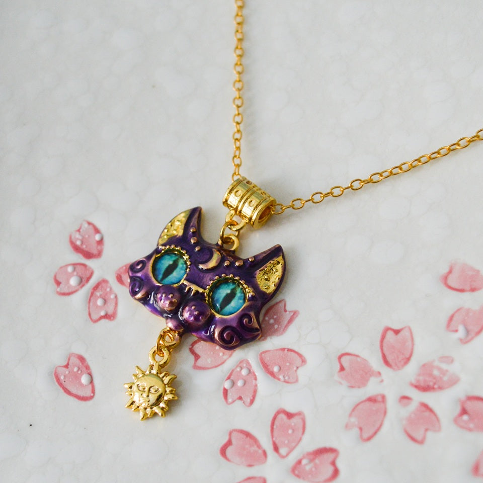 un collier doré dont le pendentif est un chat violet aux grands yeux bleus expressifs. Il a des décorations dorées comme une breloque en forme de soleil.