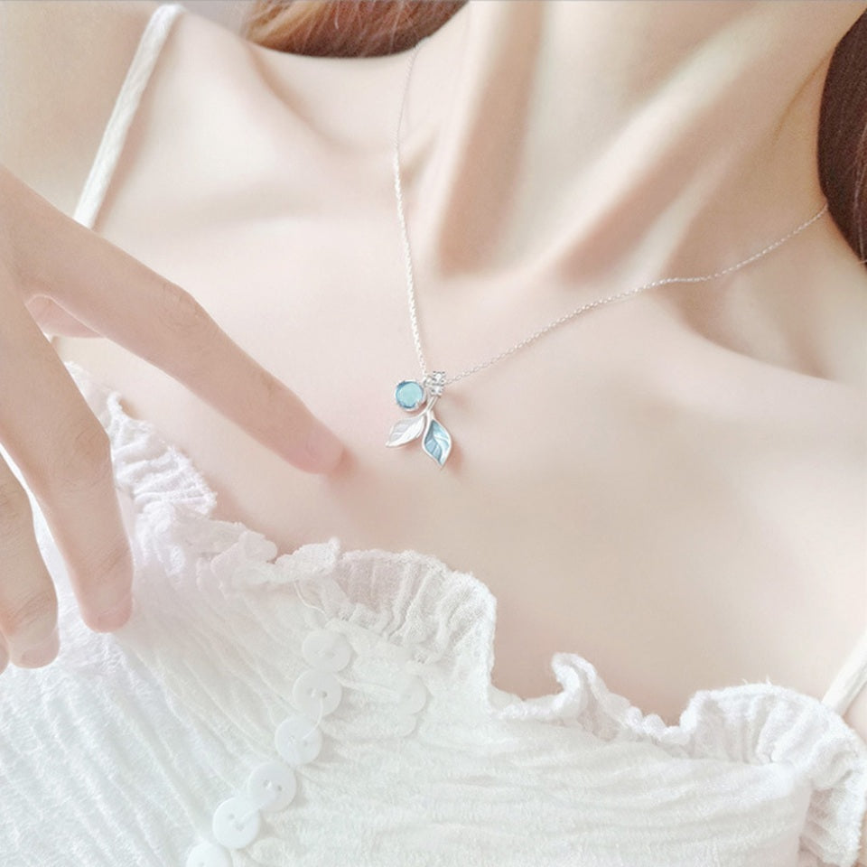 un collier argenté dont le pendentif représente de délicates feuilles bleues et d'une perle aux reflets bleutées. L'ensemble est poétique.