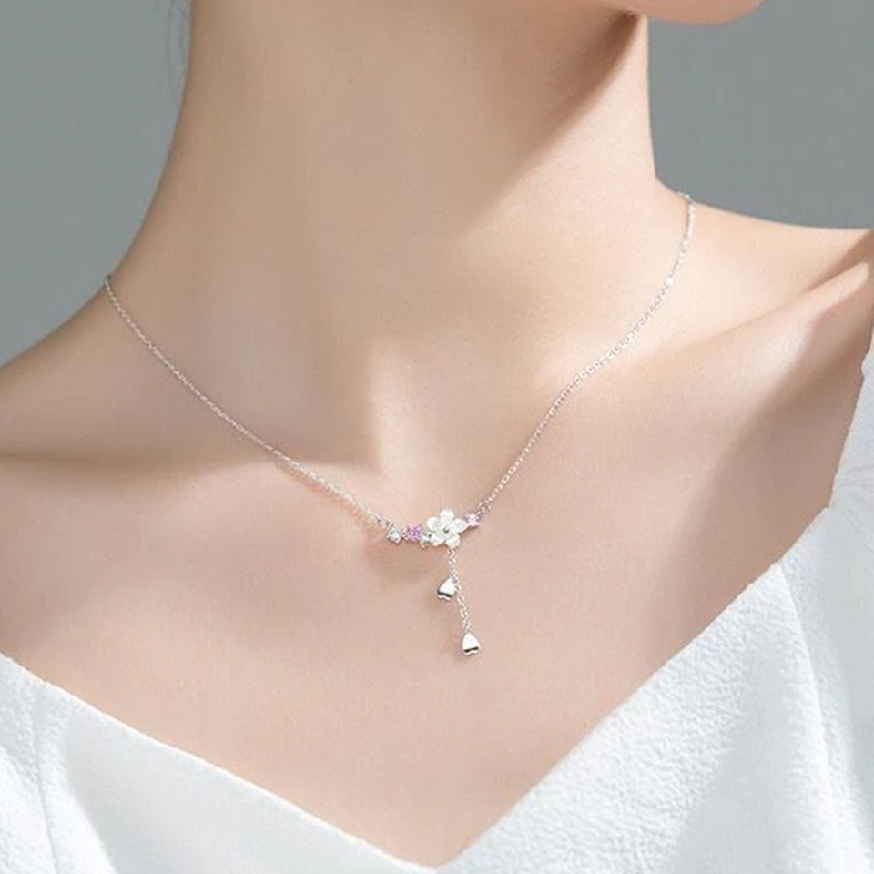 un collier argenté au design d'une délicate fleur de sakura blanche d'où pend une chainette ornée de pétales de saura. Autour de la fleur, il y a des petits strass blancs et roses qui ajoutent de la brillance.