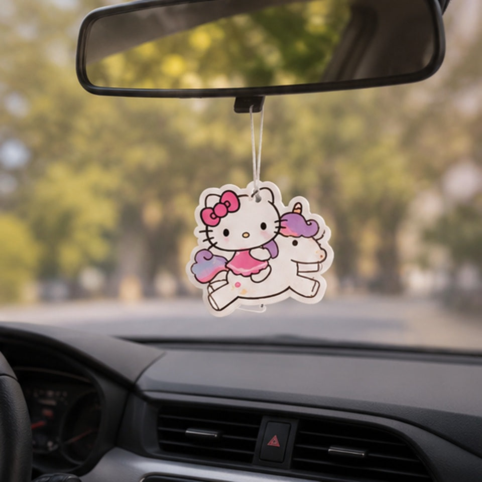 un désodorisant en forme de hello kitty sur une licorne colorée, suspendu à un rétroviseur intérieur d'une voiture, qui roule sur une route de campagne ensoleillée.
