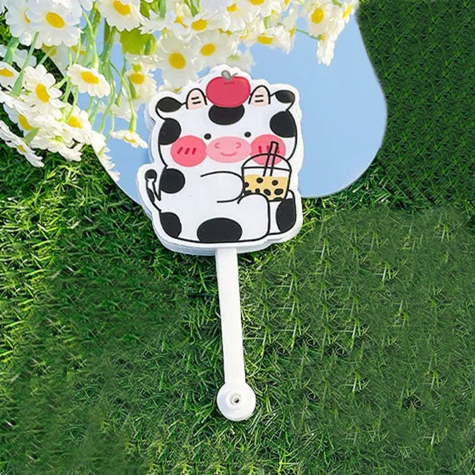 un éventail dont les lames représentent de mignonnes vaches noires et blanches qui boivent un bubble tea et qui ont une pomme rouge sur la tête. L'ensemble est adorale, c'est un accessoire utile et pratique pour les chaudes journées.