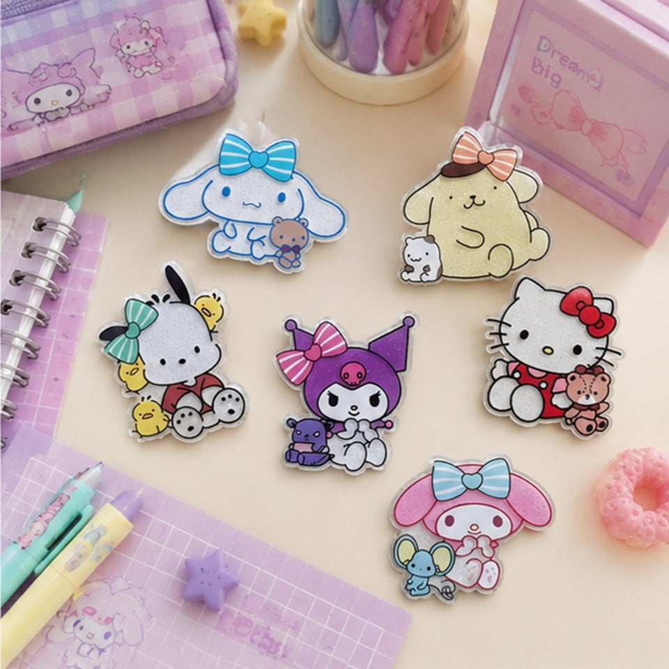 Magnets pailletés kawaii représentant plusieurs personnages Sanrio : Kuromi, cinnamoroll, Pompompurin, Hello Kitty, Pochacco et My Melody, placés sur un bureau d'écolière kawaii aux tons pastel, avec stylos roses, carnets et trousses pastels. Ambiance douce, chaleureuse et féminine.