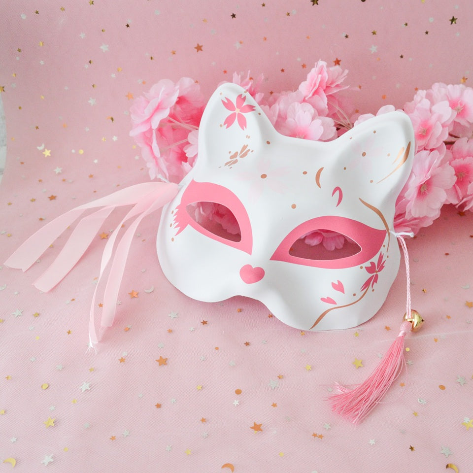 un masque kitsune rose avec des détails dorés, pompons et clochettes. Idéal pour un cosplay, une soirée déguisée ou en décoration.