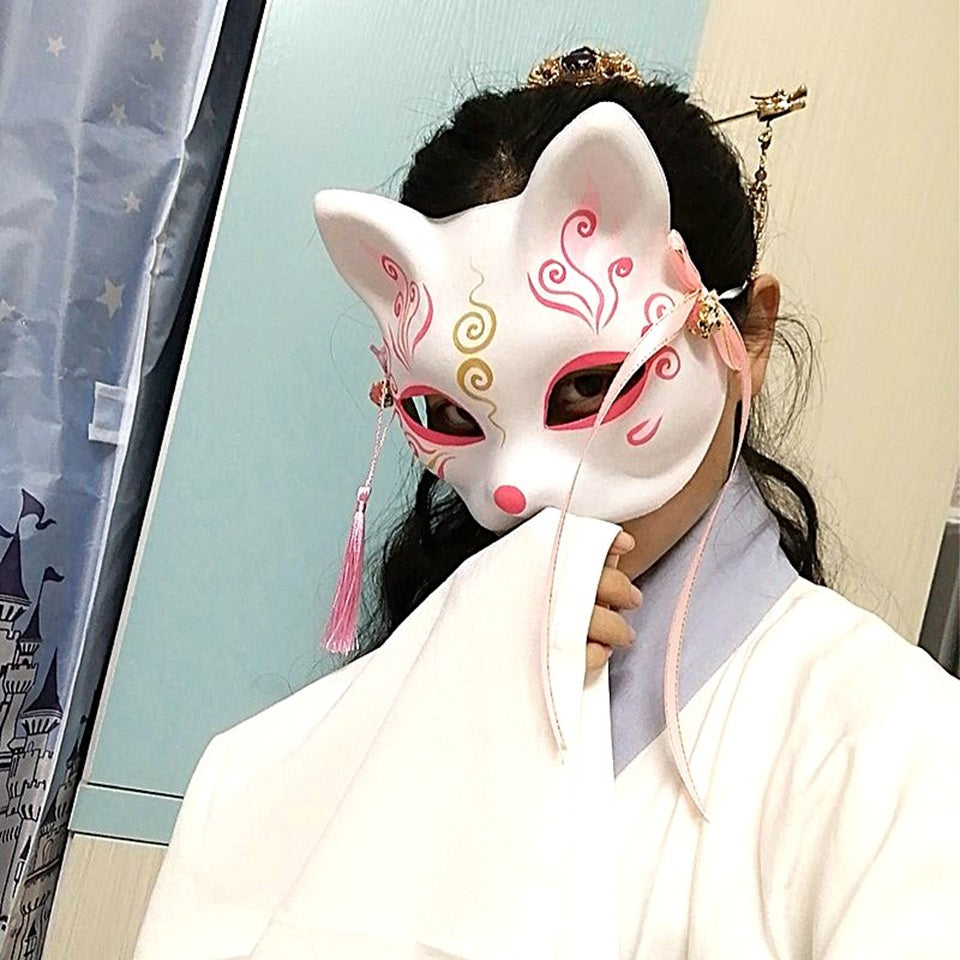 un masque kitsune rose avec des détails dorés, pompons et clochettes. Idéal pour un cosplay, une soirée déguisée ou en décoration.