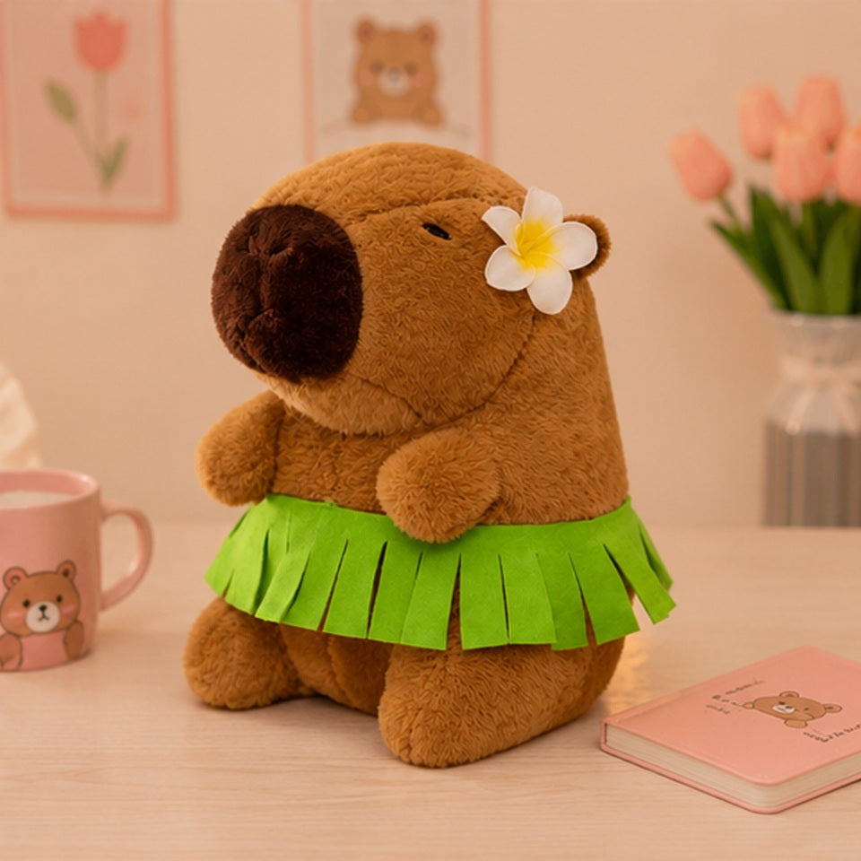 une adorable peluche marron en forme de capybara portant une jupe hawaïenne verte et une fleur blanche à l'oreille. La photo montre la peluche assise sur un lit dans une chambre kawaii aux tons pastel et chaleureux.