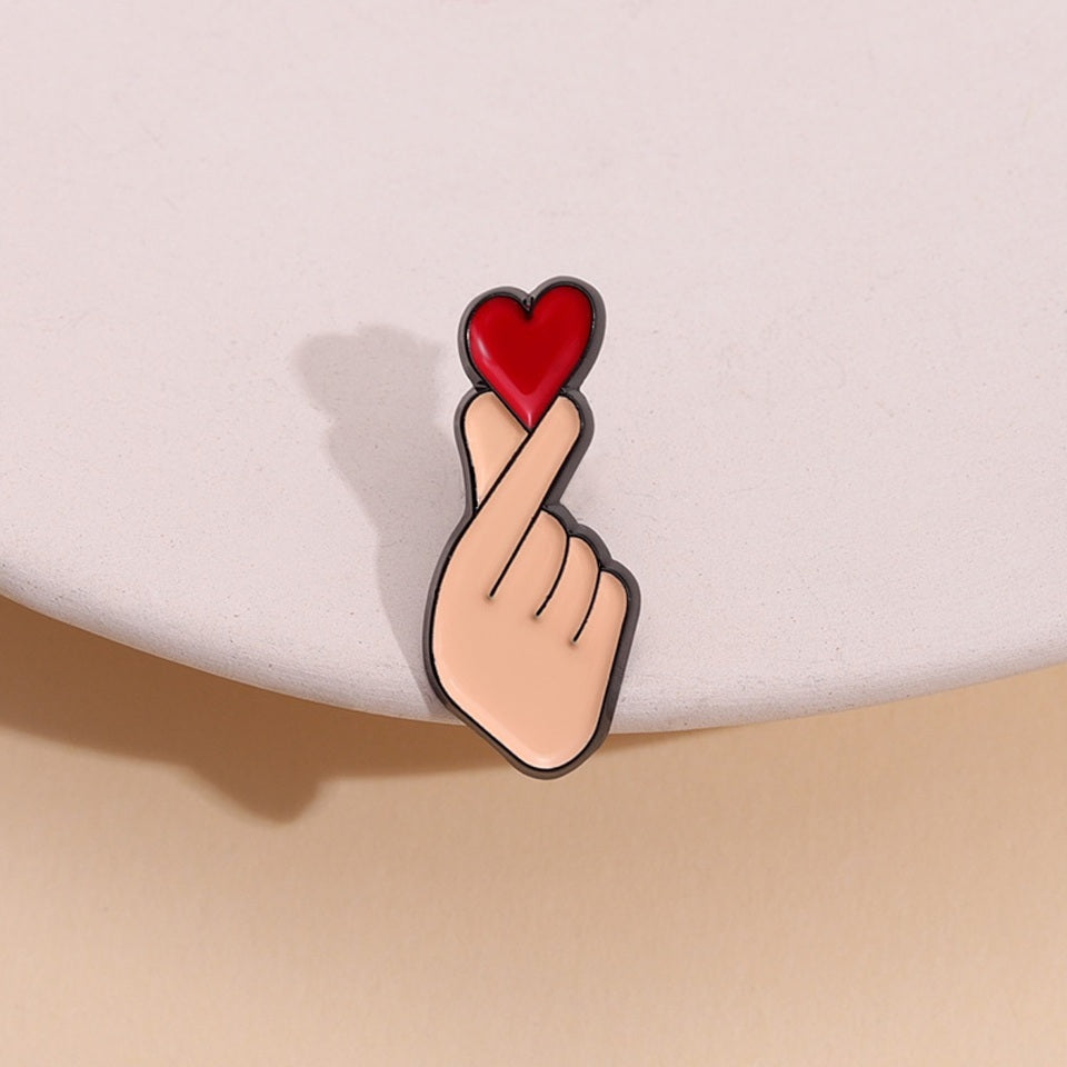 un pin's en forme de main qui forme un coeur coréen, le finger heart. Il y a un gros coeur rouge au dessus, symbole de l'amour