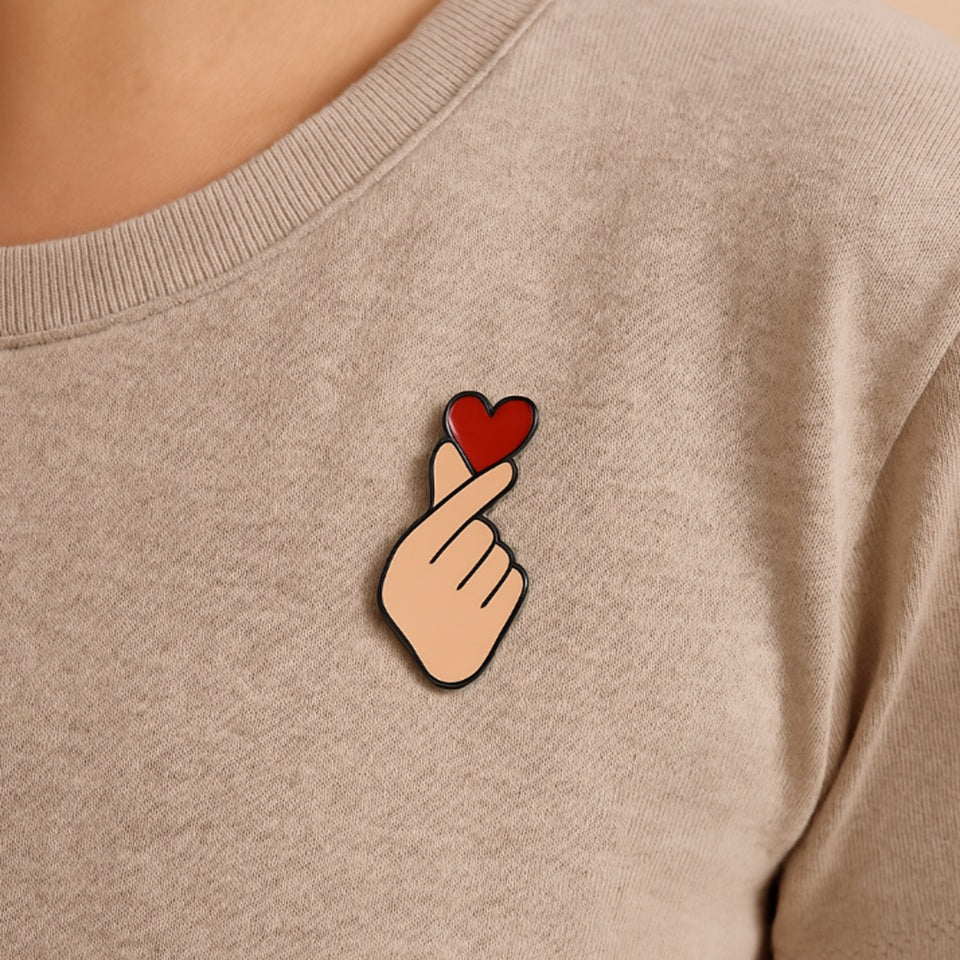 un pin's en forme de main qui forme un coeur coréen, le finger heart. Il y a un gros coeur rouge au dessus, symbole de l'amour