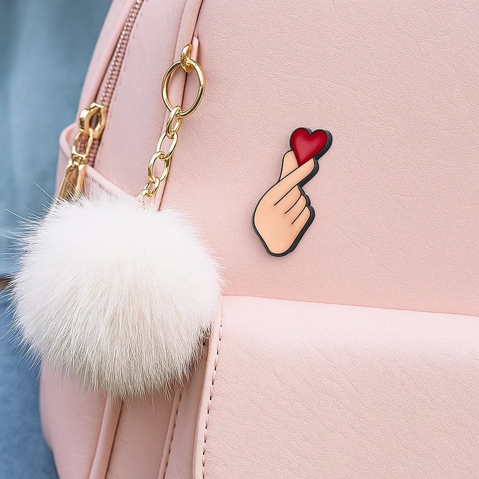 un pin's en forme de main qui forme un coeur coréen, le finger heart. Il y a un gros coeur rouge au dessus, symbole de l'amour