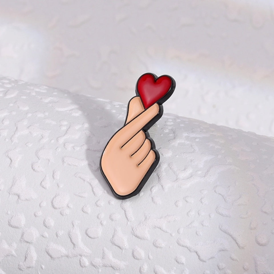 un pin's en forme de main qui forme un coeur coréen, le finger heart. Il y a un gros coeur rouge au dessus, symbole de l'amour