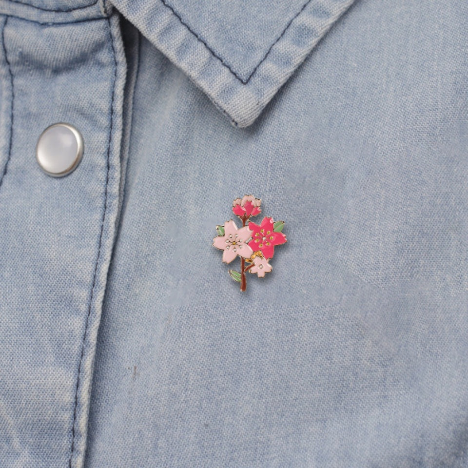 un pin's doré en forme et aux couleurs d'une branche de cerisier en fleurs. On y trouve des jolies fleurs de sakura rose foncé et rose clair et des boutons de fleurs. L'ensemble est poétique