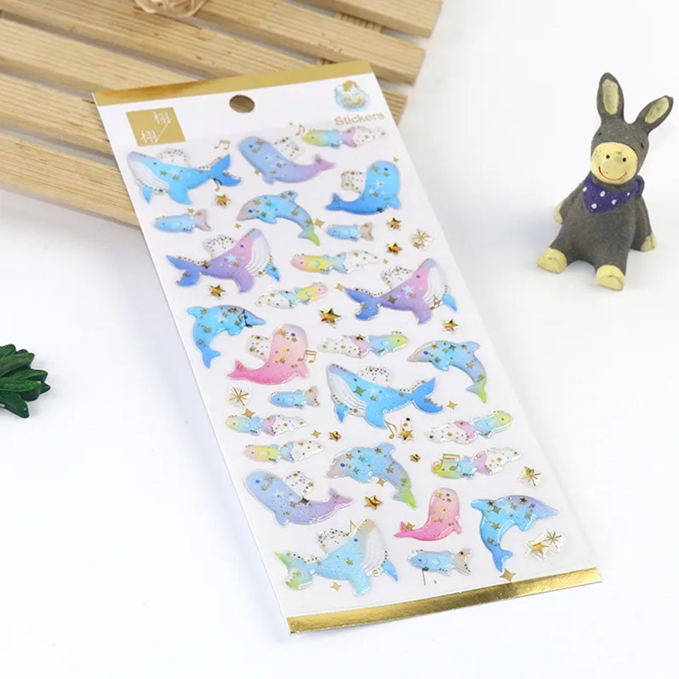 une planche de stickers en forme de baleine et de dauphin, dans les tons de couleurs bleu et violet. Ils ont des étoiles dorés qui apportent de la brillance. Effet poétique et mystérieux