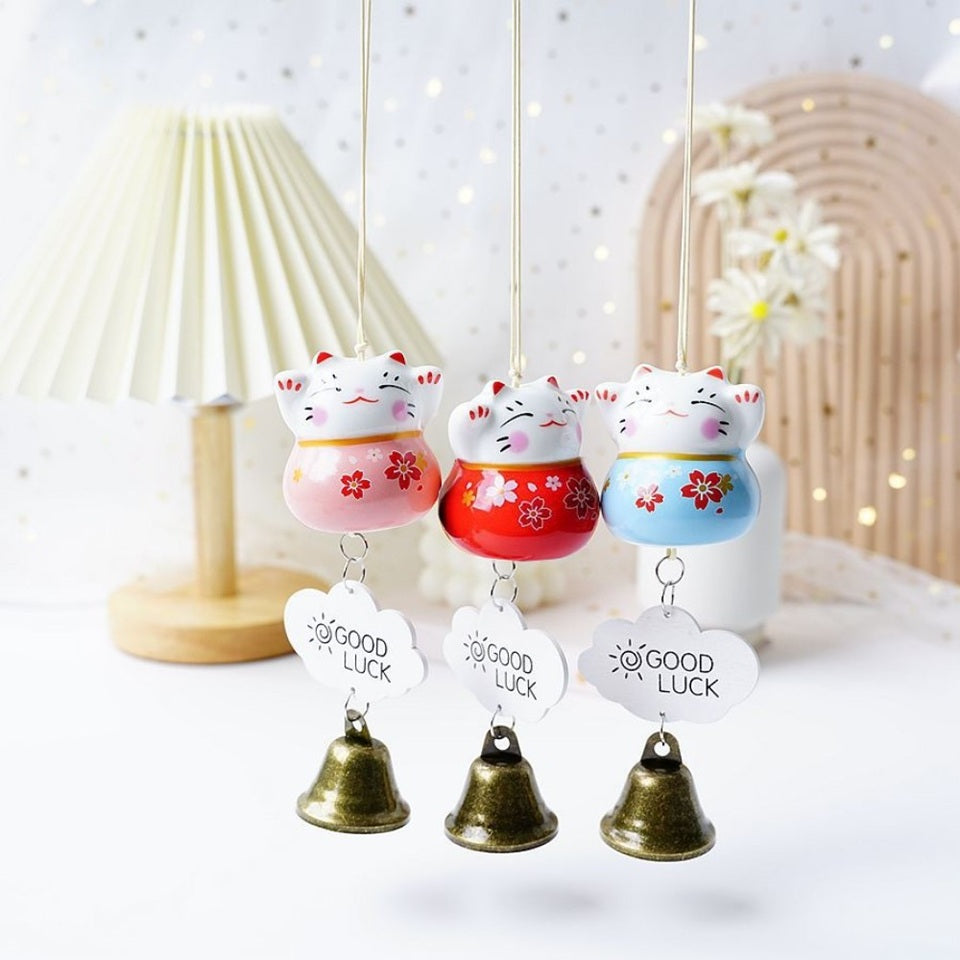 des suspensions en forme de maneki neko rouge, rose ou bleu. Une clochette y est suspendue avec une petite plaque décorative. Les maneki neko ont les pattes levées et des fleurs de sakura sur le ventre