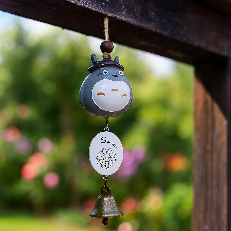 une suspension Totoro portant un petit chapeau de jardinier. une clochette et une joli epancarte en bois sont suspendus au bout. Il est très mignon et est à suspendre à un porche d'entrée ou une fenêtre