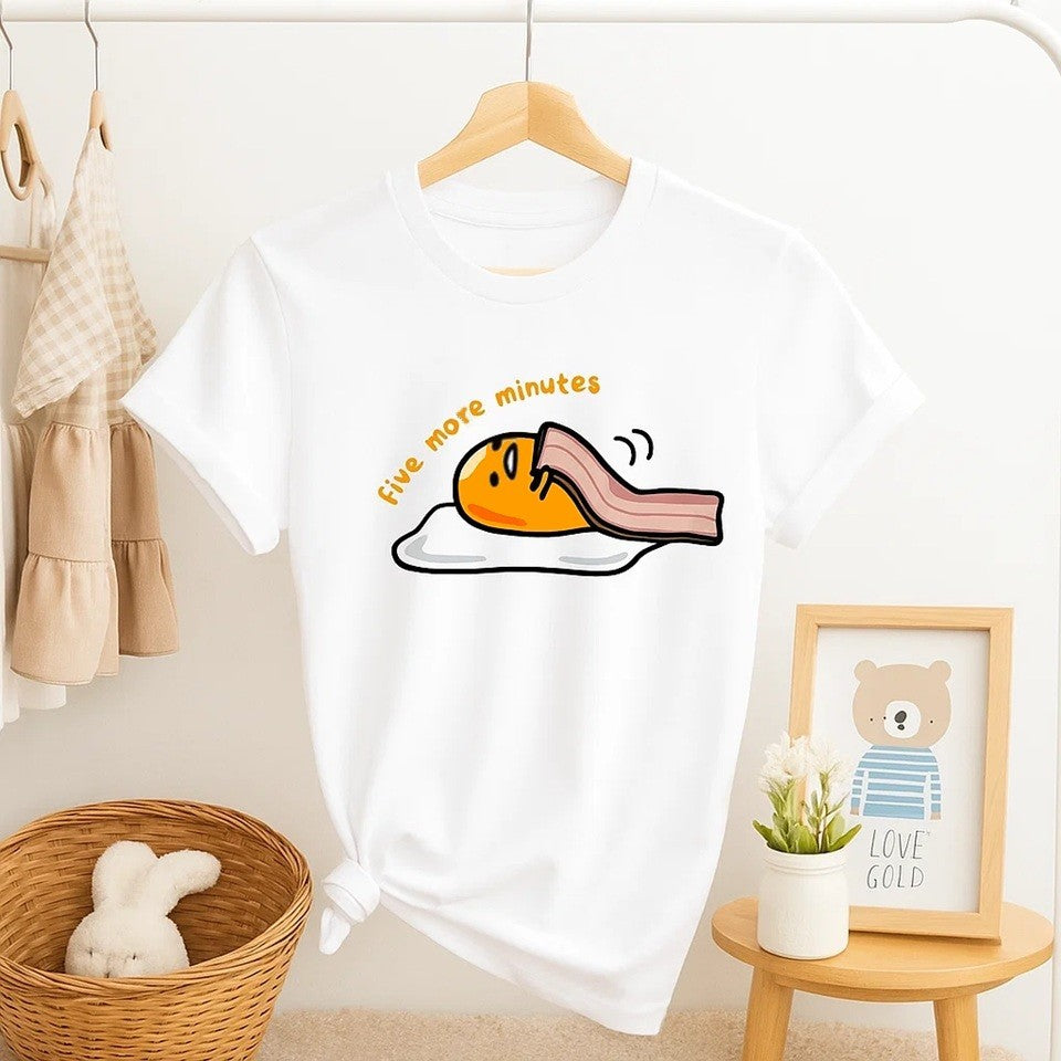 un tee-shirt blanc sur lequel est représenté Gudetama, l'oeuf mignon et rigolo de Sanrio. Il est recouvert d'une tranche de bacon comme une couverture avec le message suivant : encore 5 minutes de plus.