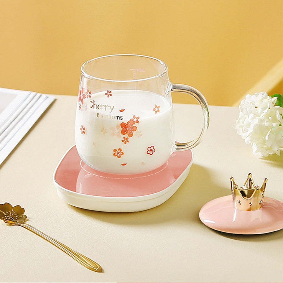 une tasse transparente au design de fleurs de sakura rose posée sur un support rose sur une table blanche. A côté, se trouve le couvercle rose orné d'une couronne dorée et la cuillère en forme de fleur de sakura. Design floral mignon et coloré qui rappelle le Japon